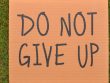 Cardboard sign reading 'Do Not Give Up' placed on green turf, symbolizing perseverance.