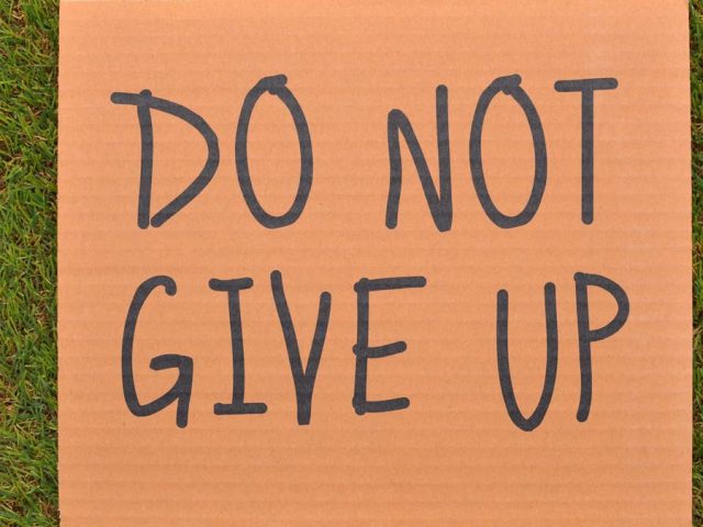 Cardboard sign reading 'Do Not Give Up' placed on green turf, symbolizing perseverance.